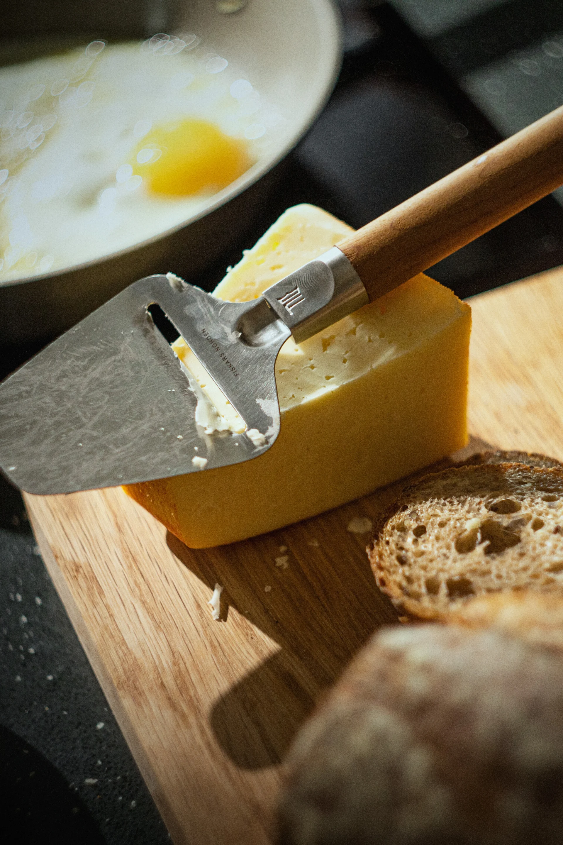 一把切芝士刀放在木板上的一块黄色芝士上，背景是面包片和煎锅里的一个煎蛋。