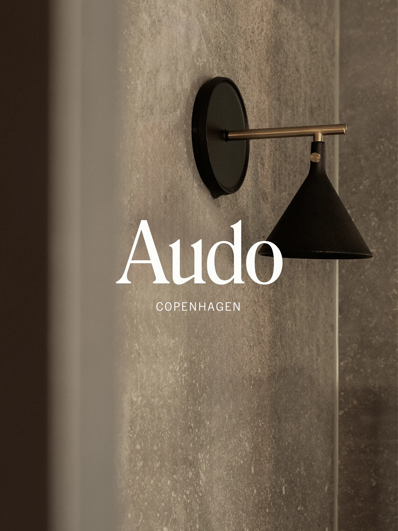 一盏壁挂式黑色灯，带黄铜臂，位于纹理灰色墙壁上，并有“Audo Copenhagen”文字。