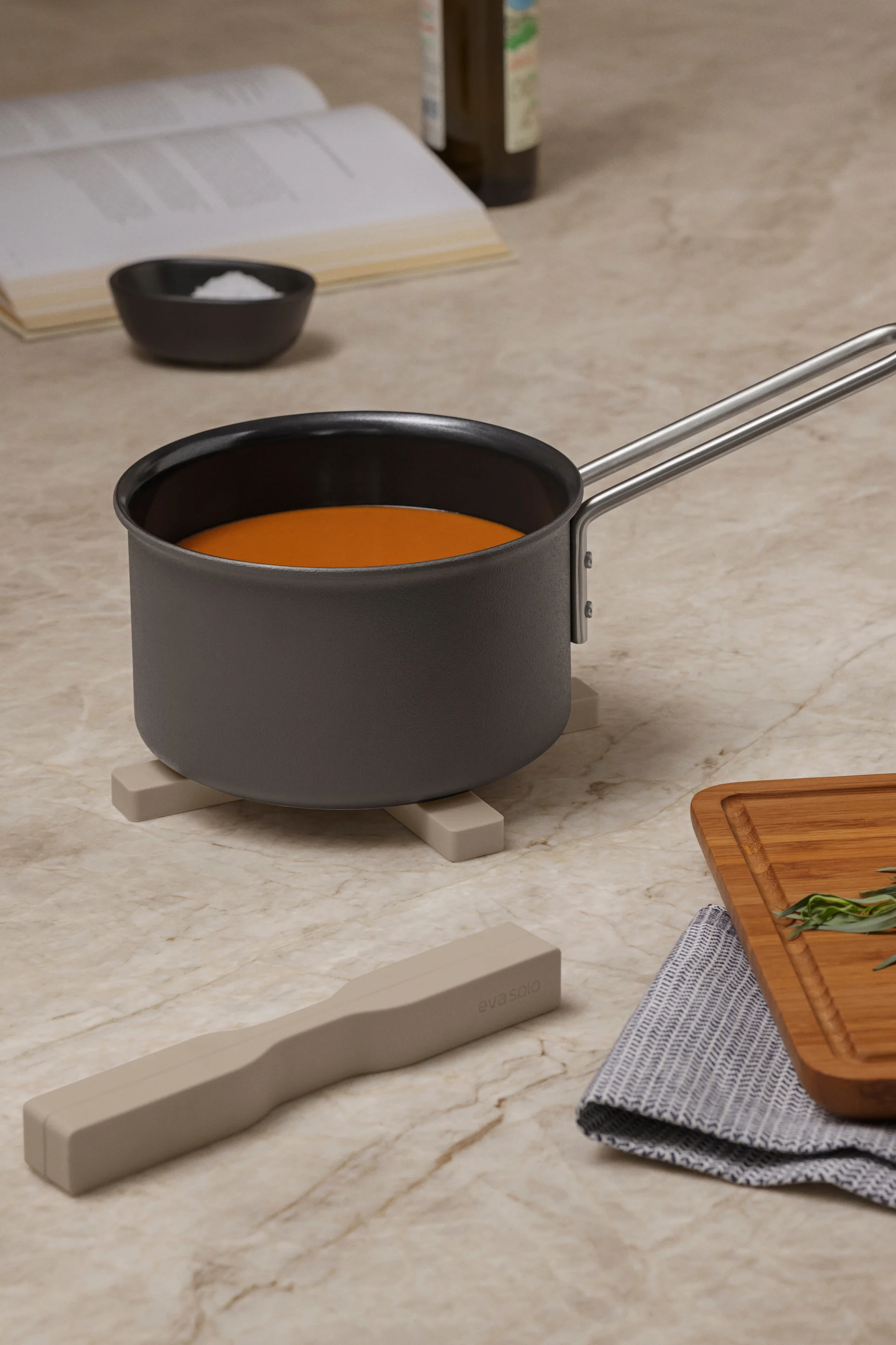 带有橙色液体的平底锅放在锅垫上，旁边有食谱、盐、打蛋器、砧板上的香草和大理石台面上的餐具。