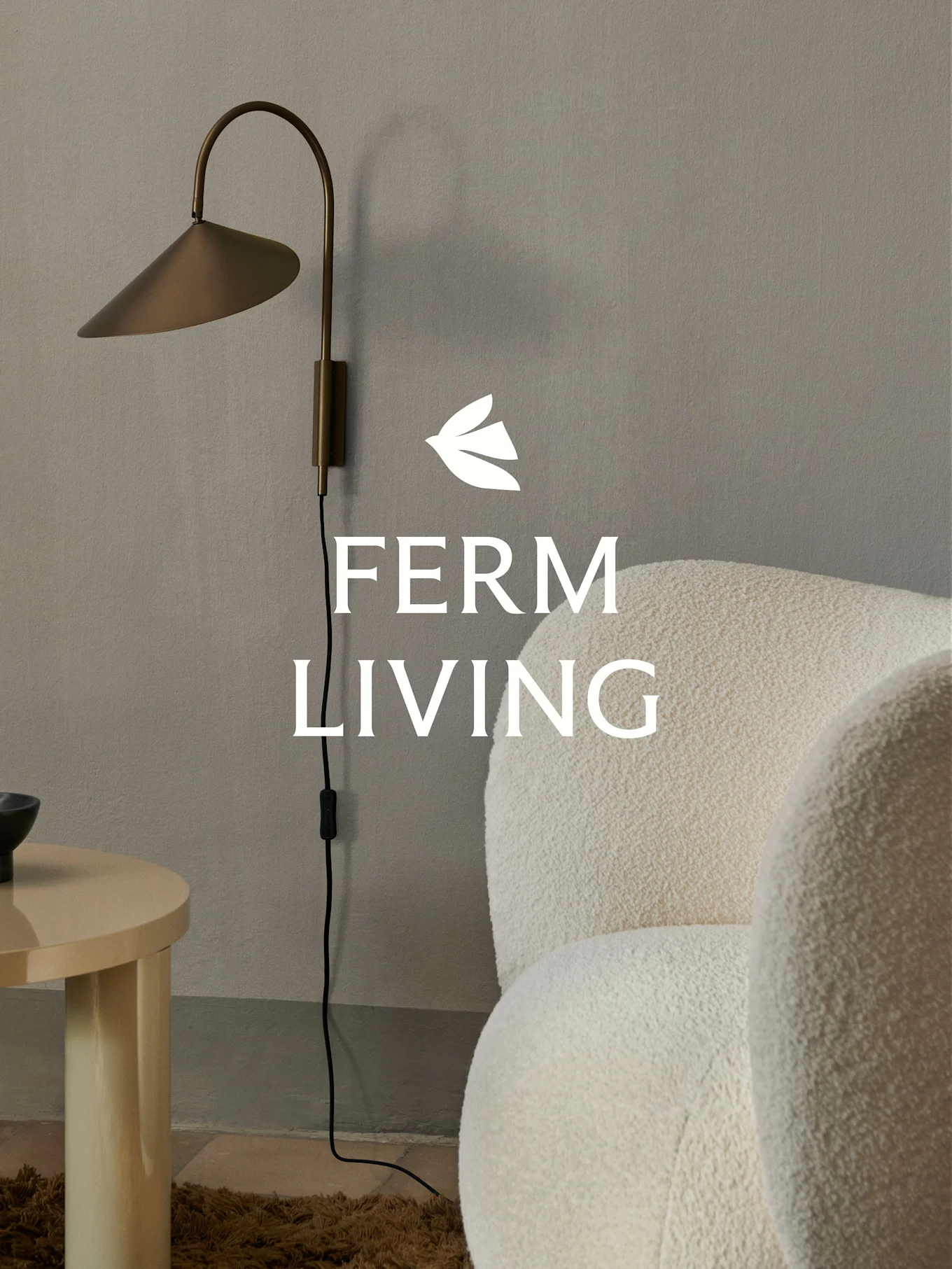 Ferm Living标志，黄铜壁灯、米色羊毛圈扶手椅和浅色木质边桌在一个简约的房间内。