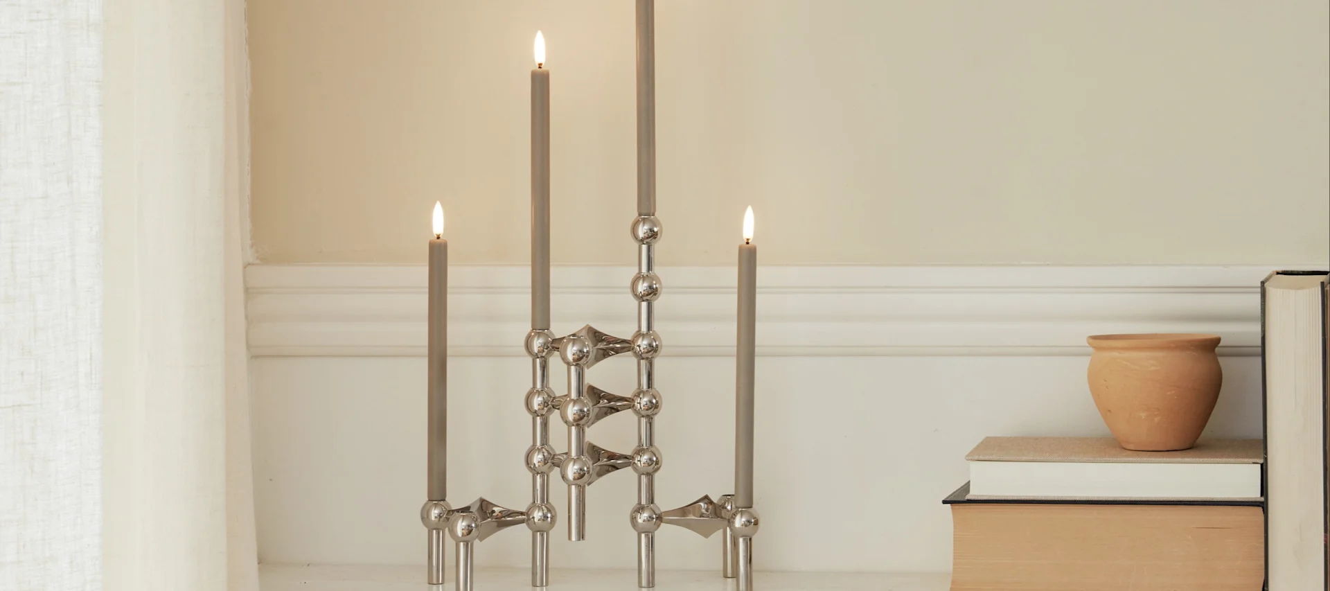 三个模块化银色烛台，上面有燃着的灰色蜡烛，旁边放着一个陶罐和书籍。