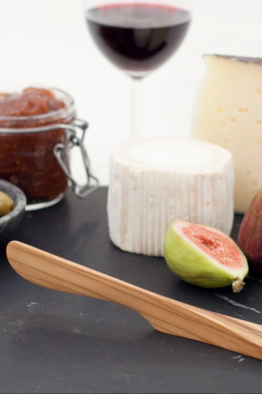 一个芝士板，上面有木制刀、两种芝士、半个无花果、果酱、橄榄和一杯红酒。