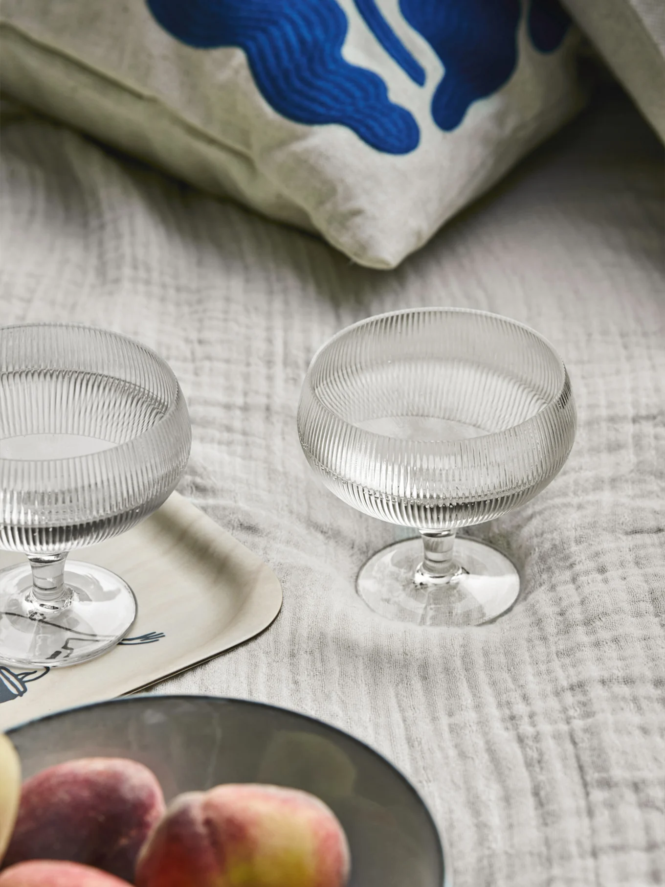 两个带罗纹的玻璃甜点碗、一个带有蓝色抽象艺术图案的枕头、一个木托盘和浅色织物上的桃子.